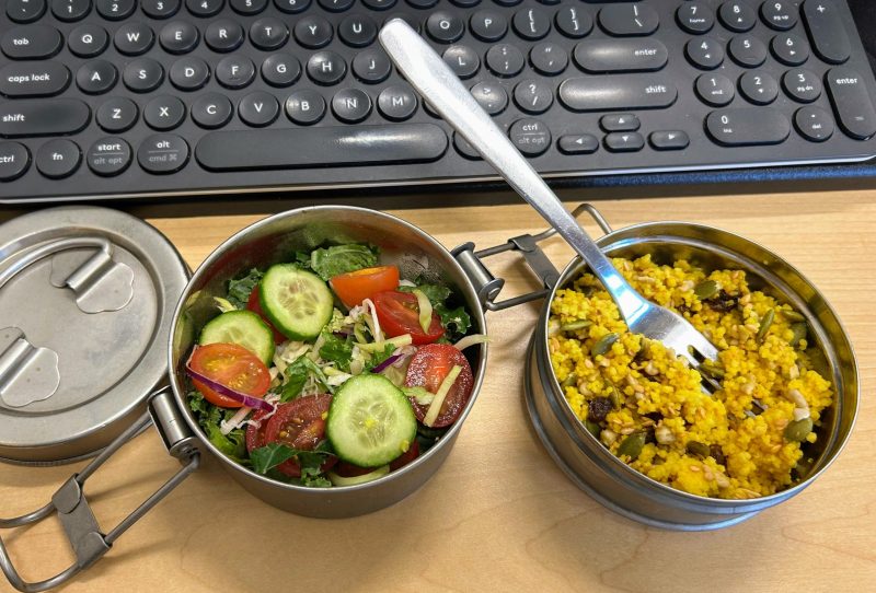 Two steel containers and a lid next to a keyboard on a desk. One container of cous cous and another of salad.